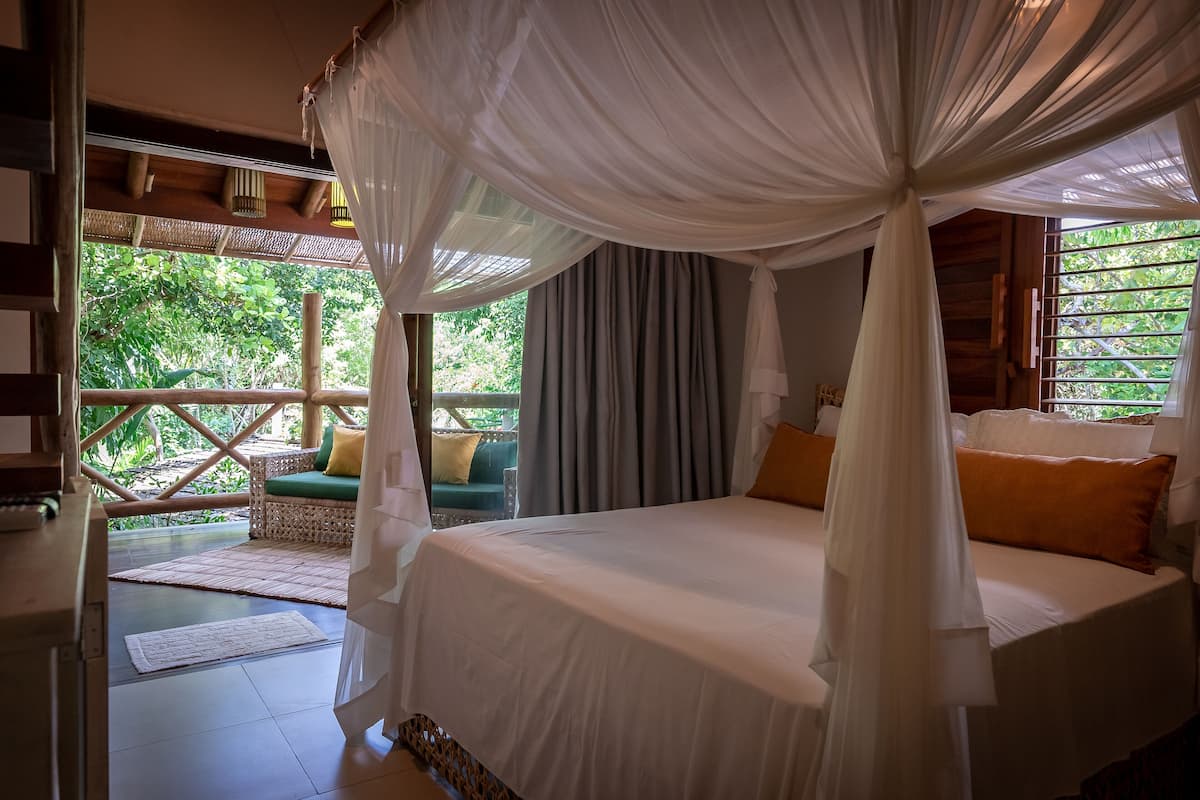 Ein Schlafzimmer mit Baldachinbett, Blick auf Grünflächen und einem Korbssessel mit Kissen.