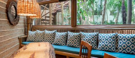 A wooden bench with patterned cushions, a woven lamp, and a view of tropical plants through large windows.