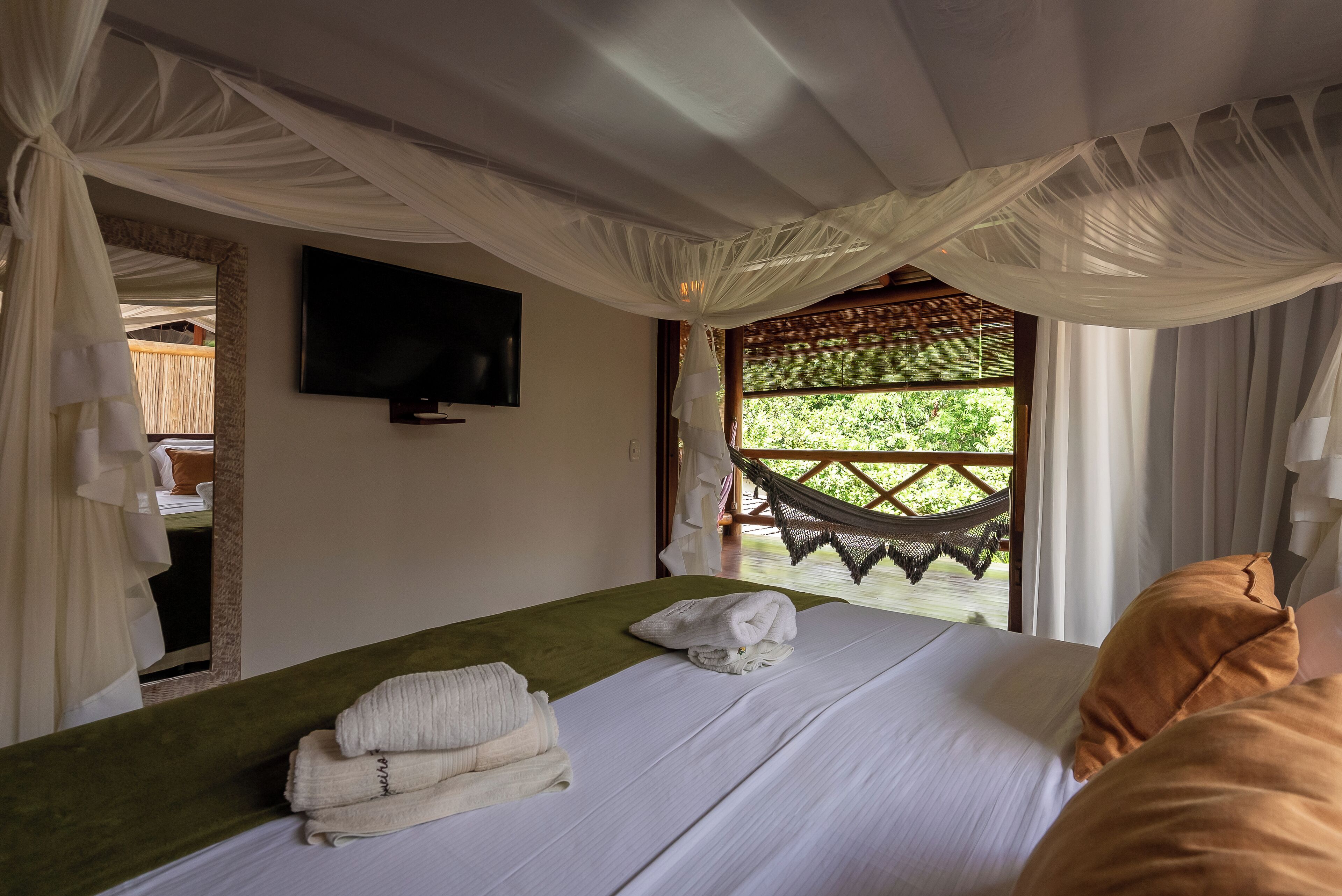 Quarto com cama, televisão, rede do lado de fora da janela e vista para o verde.