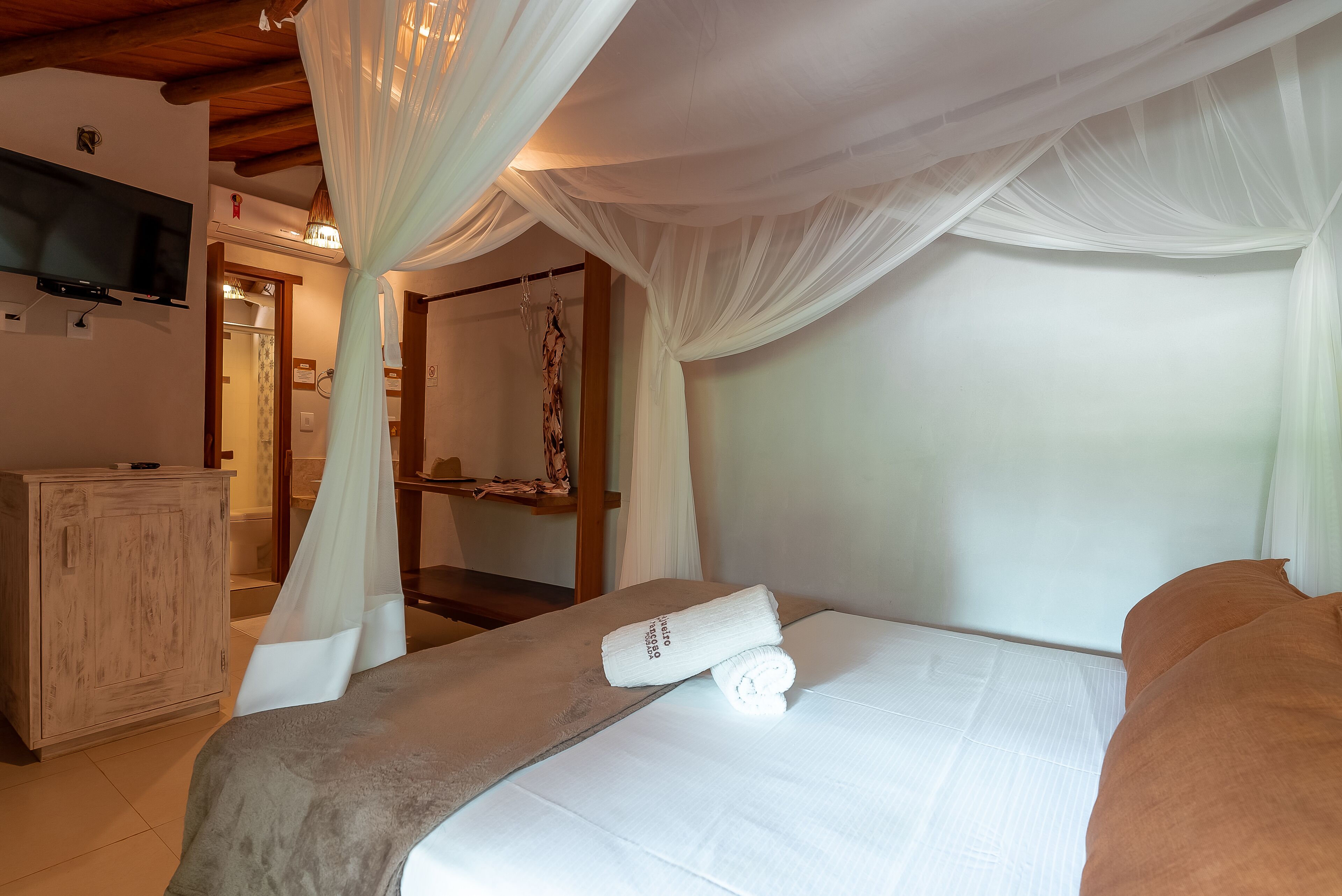 A bedroom with a bed, a wooden dresser, a television mounted on the wall, and a bathroom visible in the background.