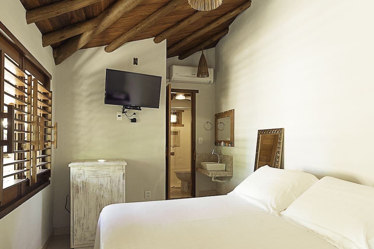 Ein Schlafzimmer mit Holzbalkendecke, einem Bett, einem Fernseher, einem Badezimmer und einem Fenster mit Jalousien.