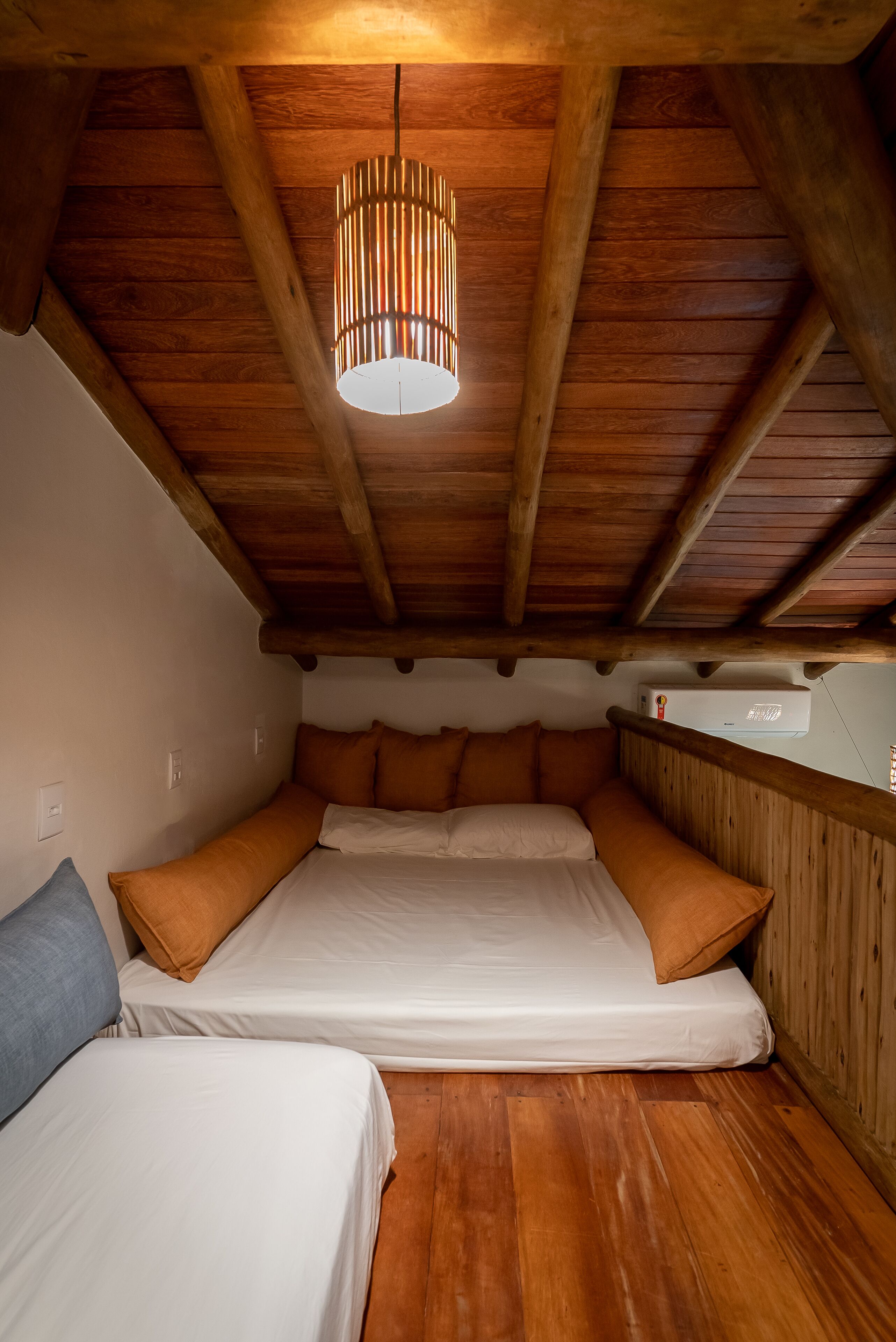 Une chambre confortable située dans les combles, avec un plafond en bois, un luminaire suspendu et un lit recouvert de draps blancs et agrémenté d’oreillers orange.