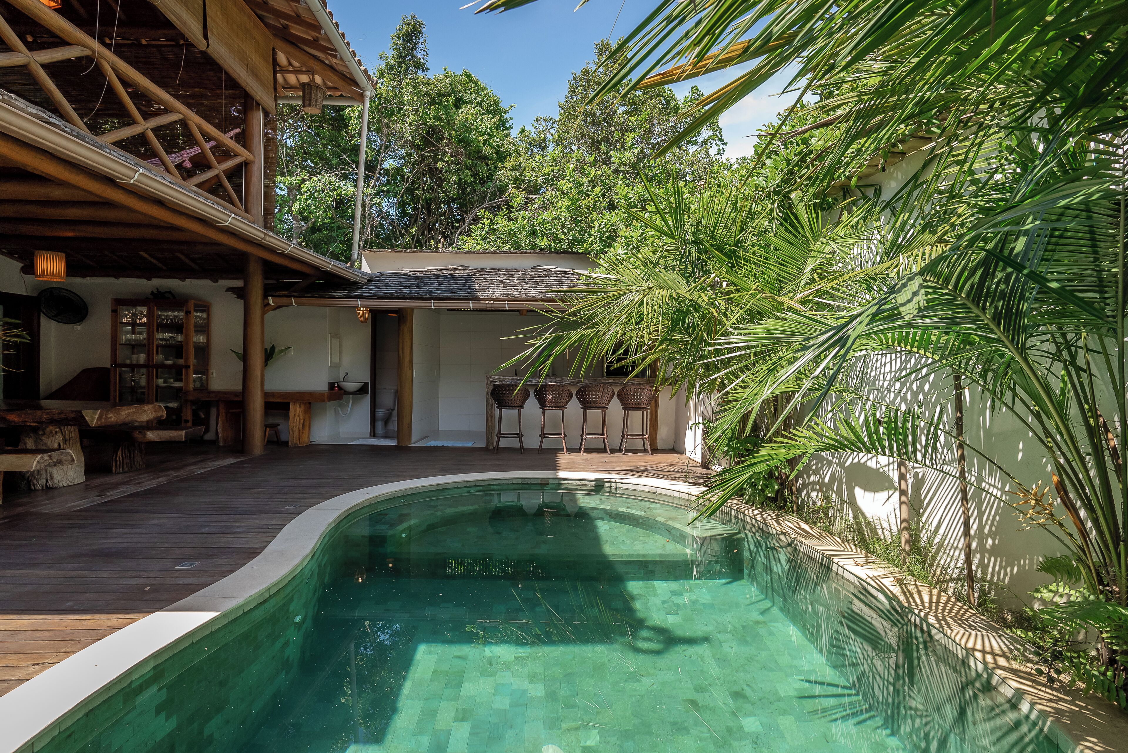 Un área de piscina con terraza de madera, zona de estar cubierta y exuberante vegetación.