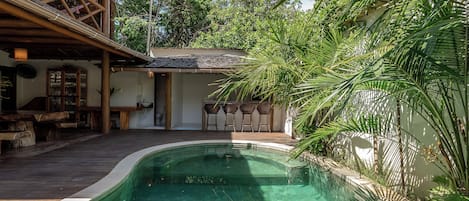 Un área de piscina con terraza de madera, zona de estar cubierta y exuberante vegetación.