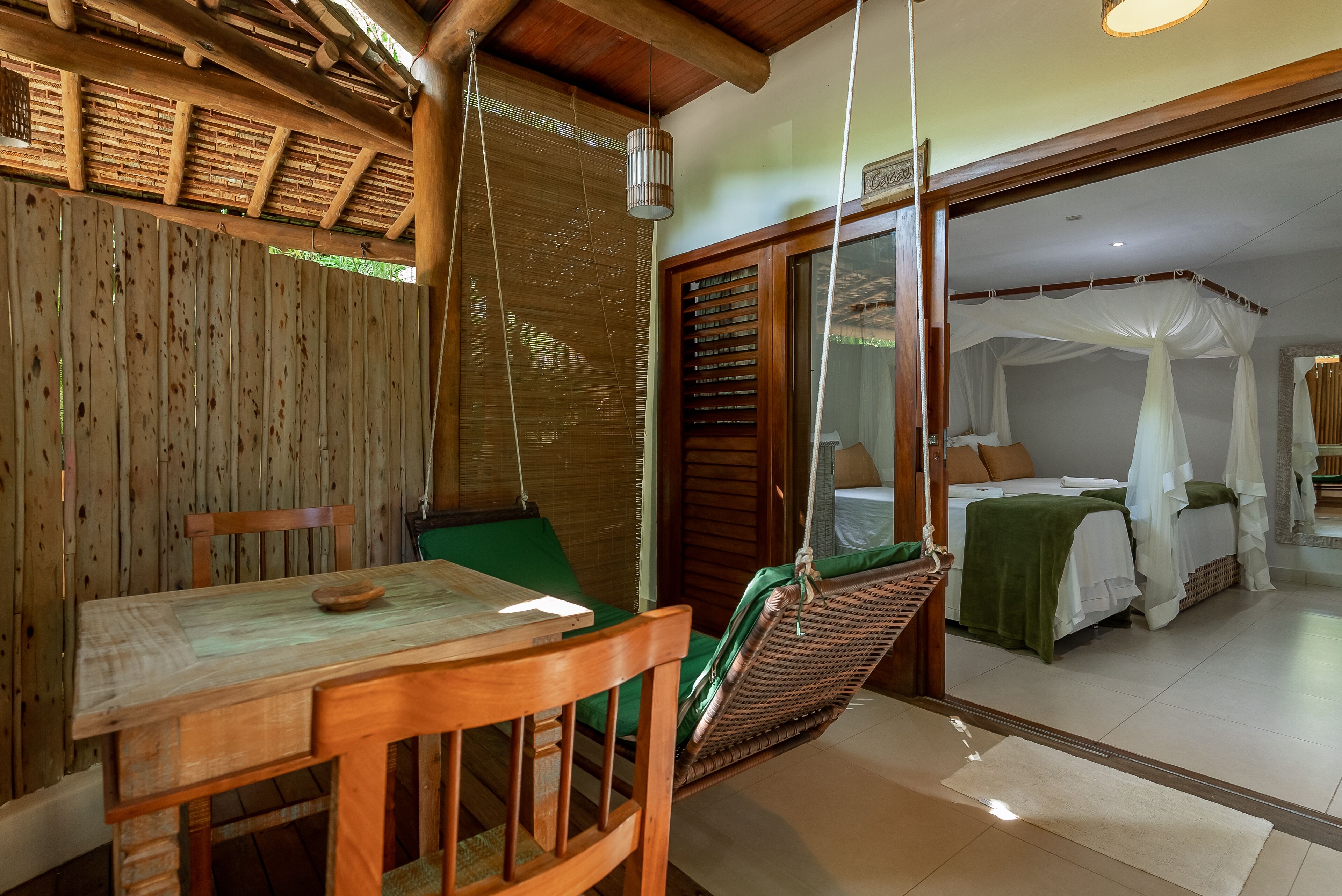Une table à manger en bois, une chaise verte, un fauteuil suspendu en osier et une chambre avec un lit placé sous un dais blanc.