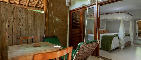 Une table à manger en bois, une chaise verte, un fauteuil suspendu en osier et une chambre avec un lit placé sous un dais blanc.
