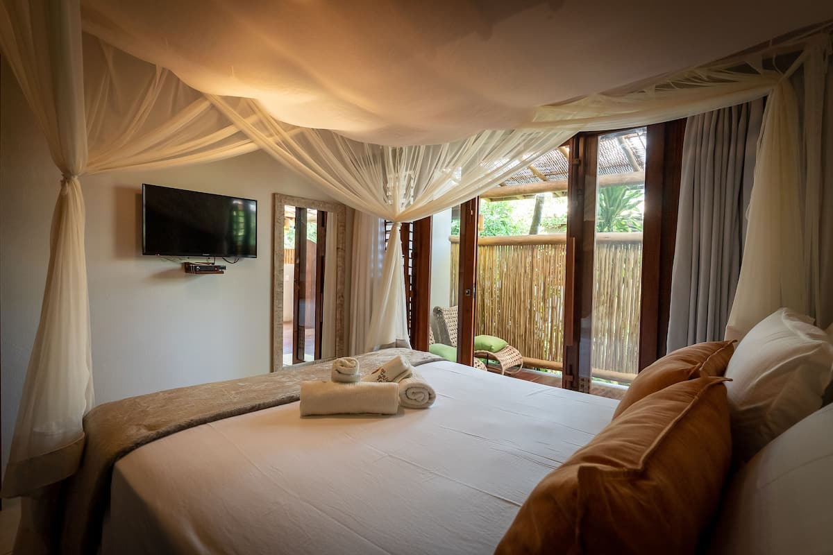Ein Schlafzimmer mit einem großen Bett, einem flachbildfernseher an der Wand und Blick ins Freie durch große Fenster.