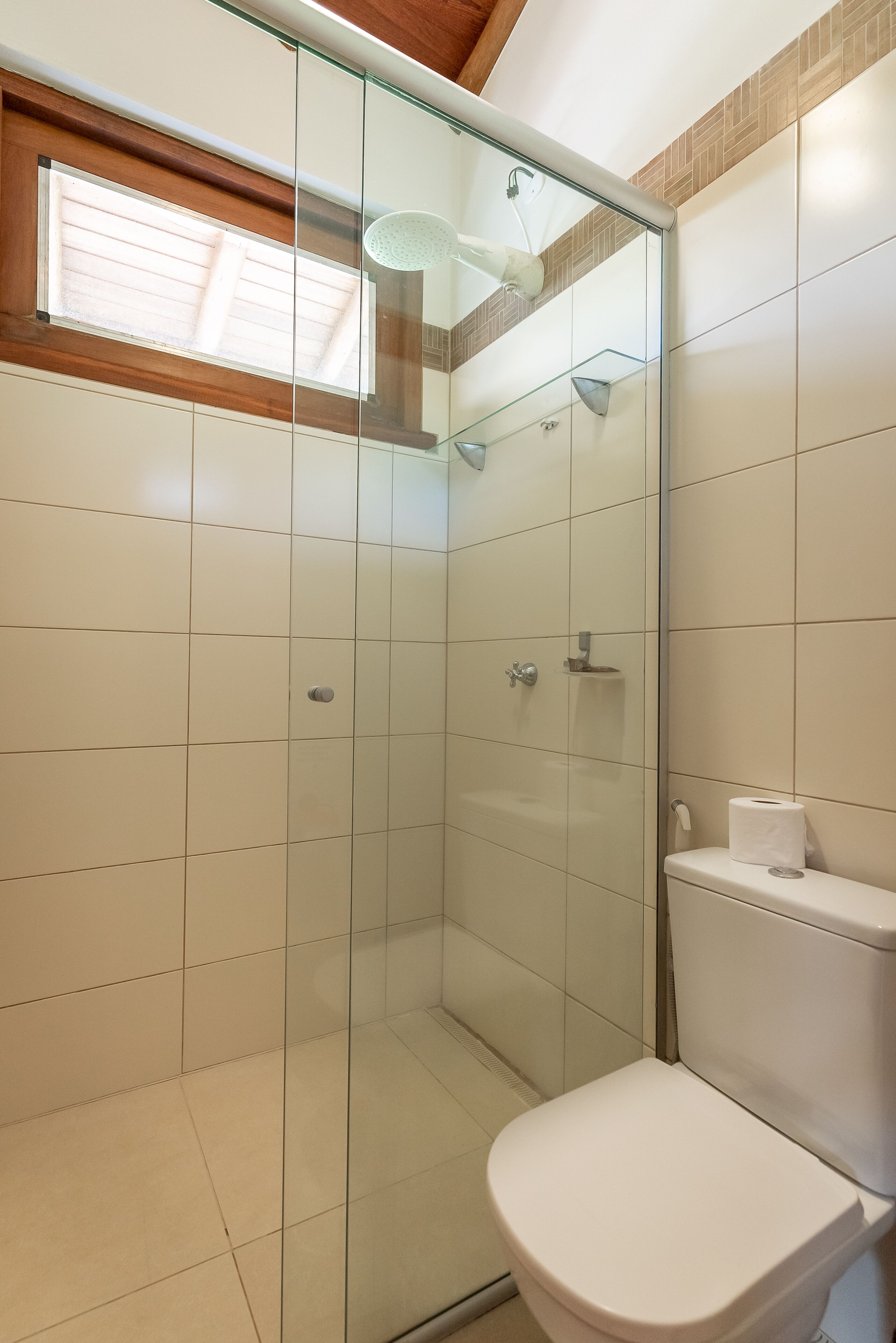 Une douche fermée par des parois de verre, avec des toilettes blanches et une fenêtre au-dessus.