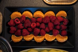 Un plateau rectangulaire noir sur lequel sont disposées, de manière ordonnée, des tranches d’orange et des framboises, le tout posé sur une surface sombre.