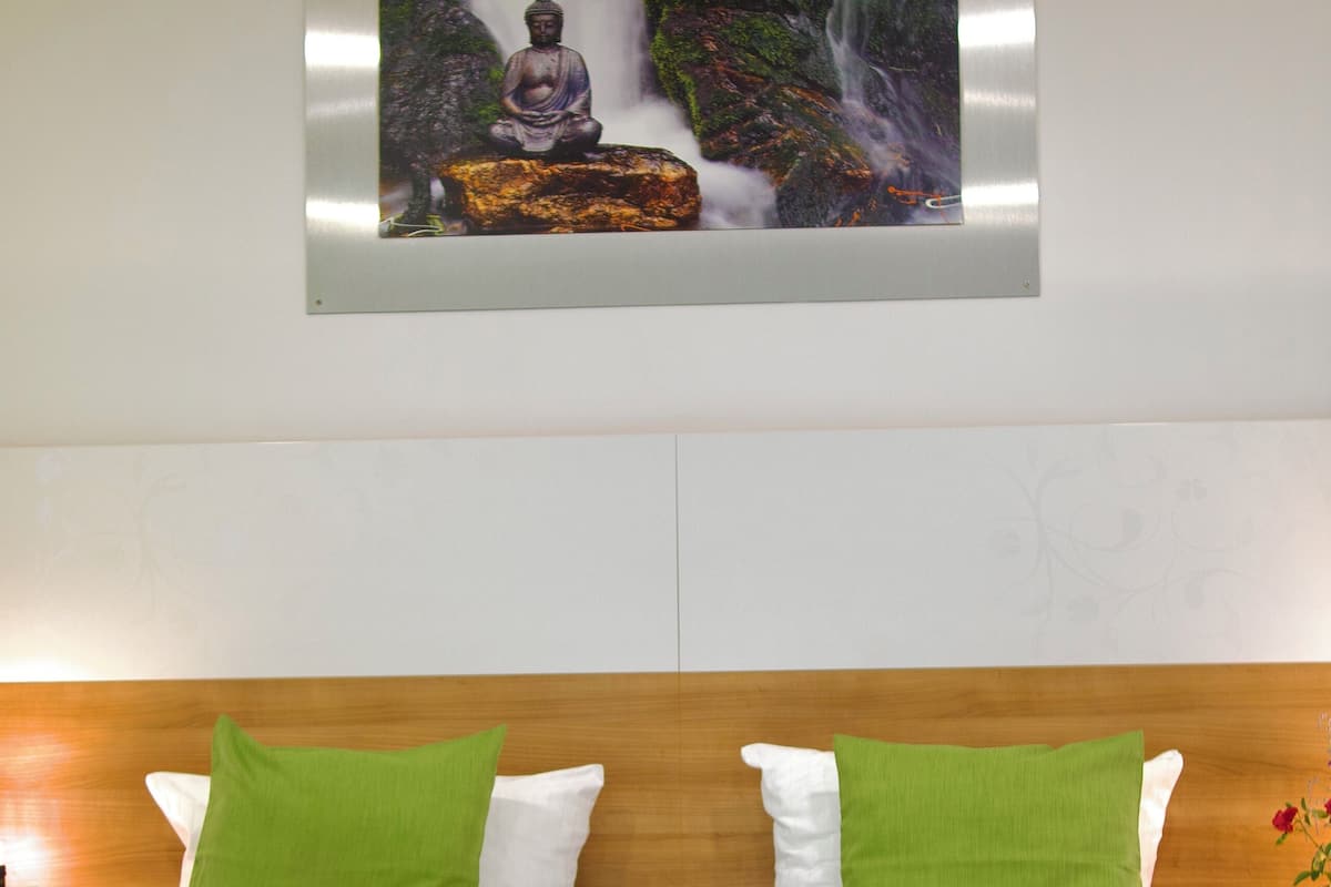 Une chambre d’hôtel avec une tête de lit en bois, des oreillers verts et blancs, un lit avec une literie verte et blanche, et un tableau encadré représentant une statue de Bouddha devant une cascade.