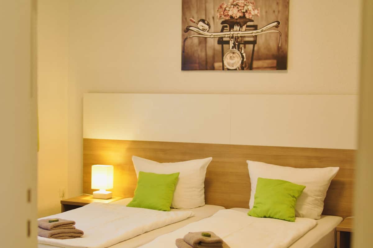 Une chambre d’hôtel avec deux lits simples, chacun doté d’une tête de lit en bois, de draps blancs et d’oreillers verts. Il y a une table de chevet avec une lampe et une serviette pliée sur chaque lit.