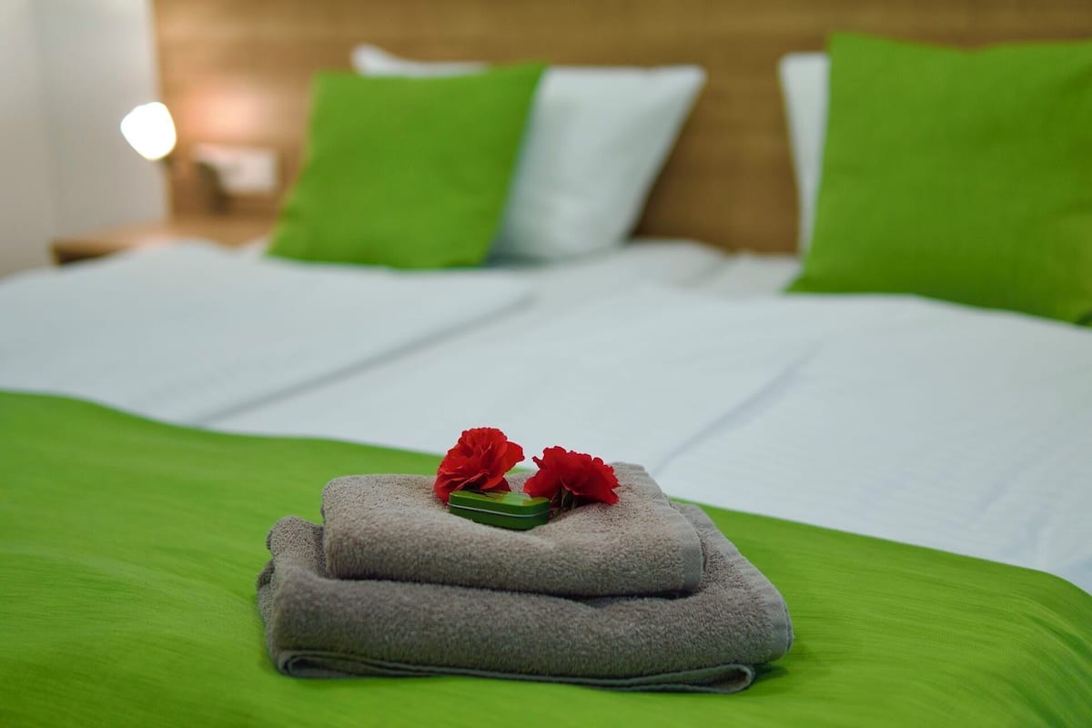 Un lit bien fait, avec une literie verte et blanche, deux fleurs rouges posées sur une serviette pliée, et une table de chevet avec une lampe en arrière-plan.