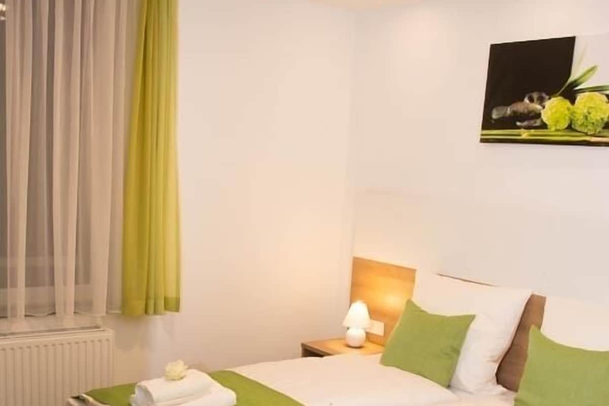 Ein Doppelbett mit grüner Bettwäsche, eine Nachttischlampe und ein Fenster mit durchsichtigen Vorhängen.