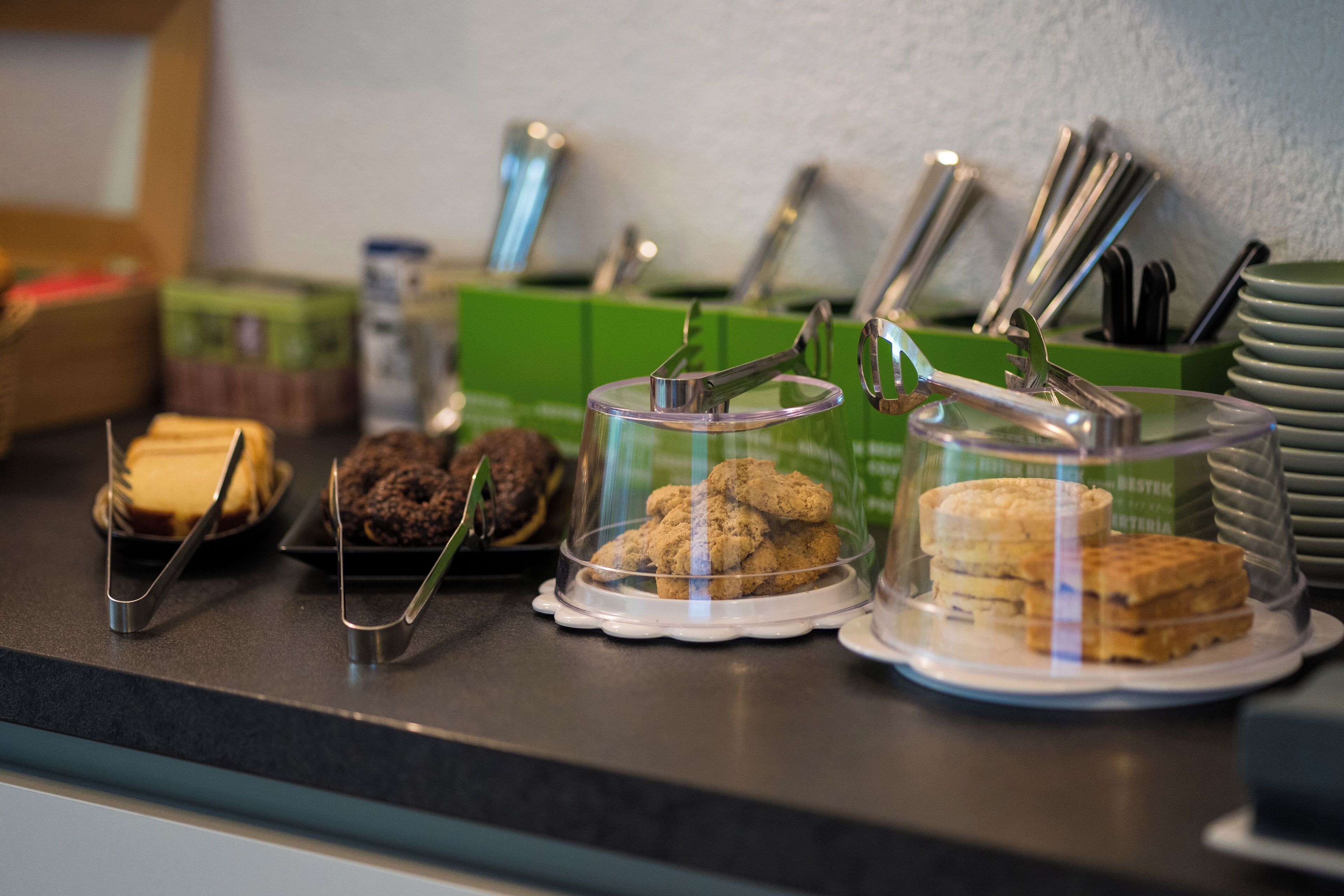 A buffet counter with various baked goods displayed in clear containers, accompanied by serving utensils and stacked plates.