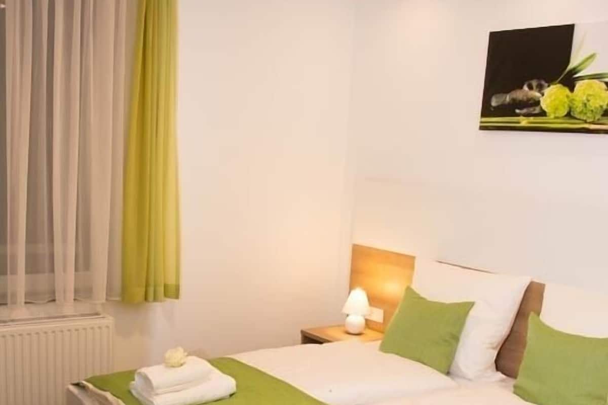 Un lit double avec une literie verte, une lampe de chevet et une fenêtre ornée de rideaux légers.