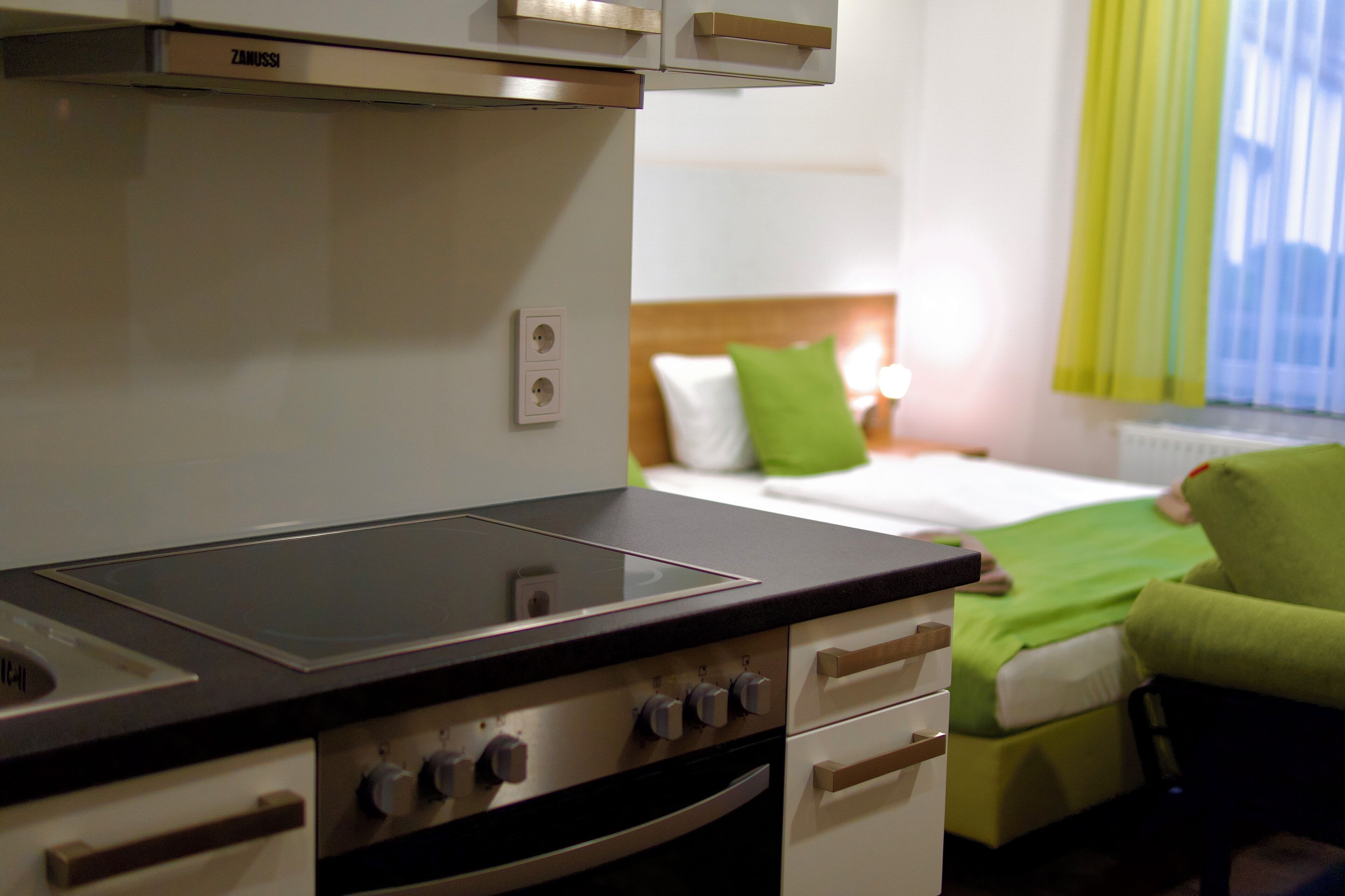 A kitchenette with a stove, oven, and two electrical outlets. A bed with a green pillow and a green blanket is visible in the background.
