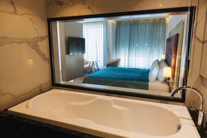 Een moderne badkamer met een groot bad, een spiegel die een slaapkamer met een bed en gordijnen reflecteert, en marmeren wanden.