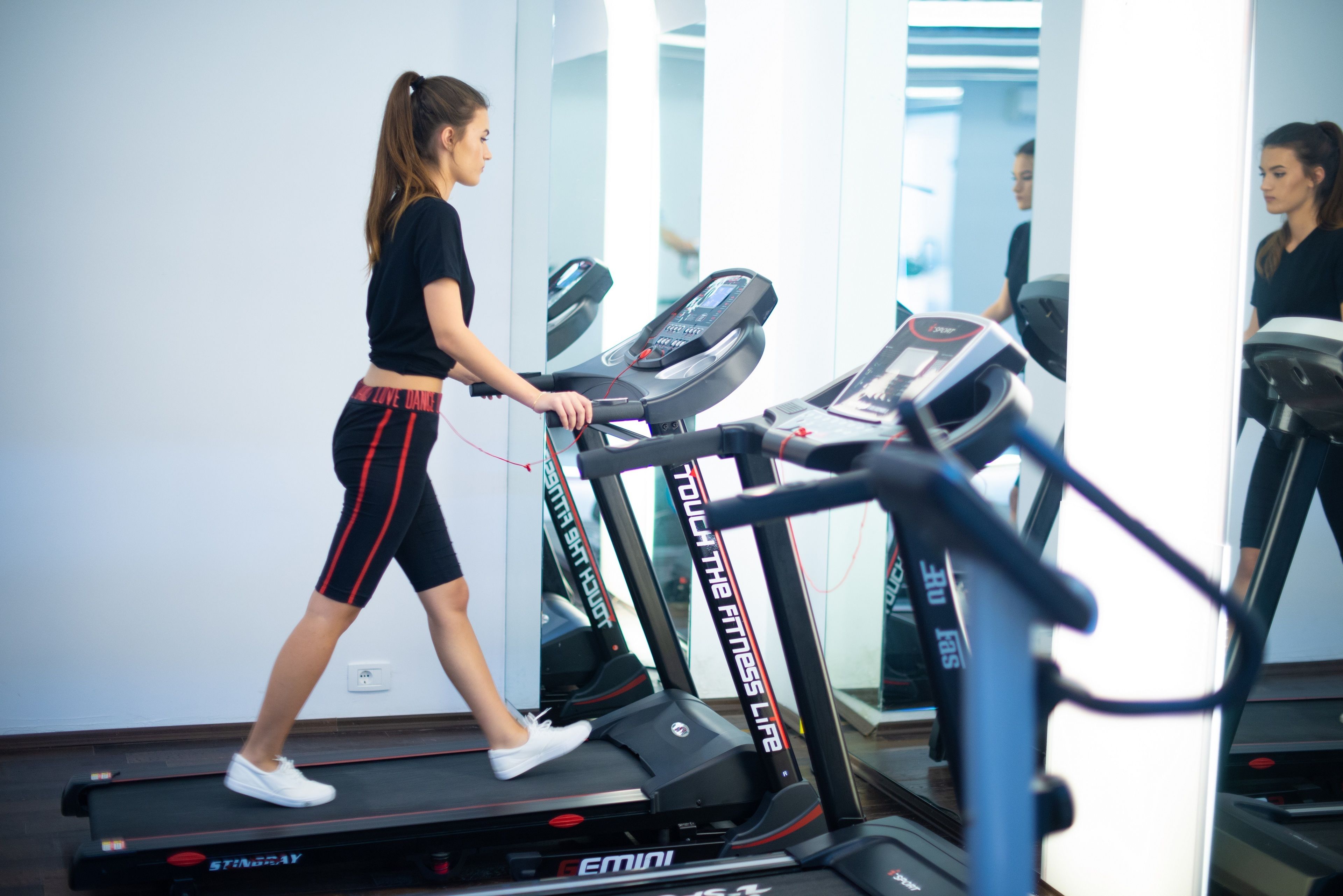 Una mujer caminando sobre una caminadora en un gimnasio con paredes espejadas y varias caminadoras.