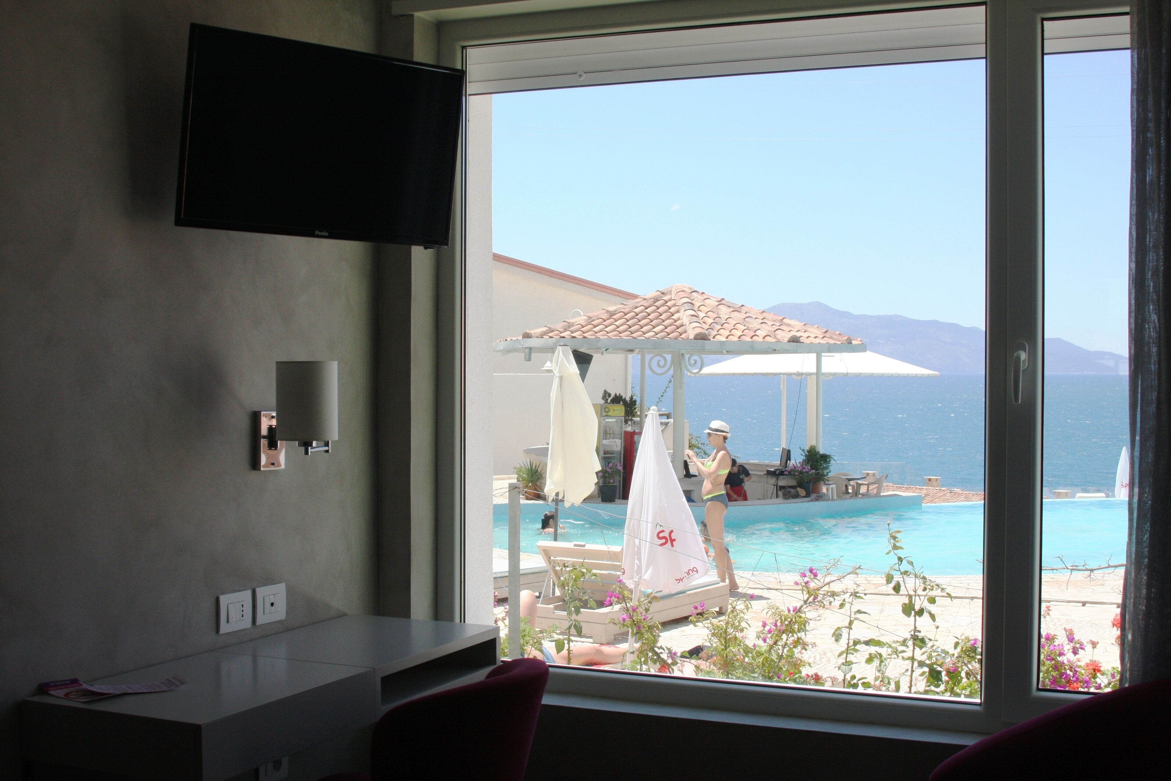 Une chambre avec un téléviseur fixé au mur, une fenêtre donnant sur une piscine et la mer, ainsi qu’une petite table avec une chaise.