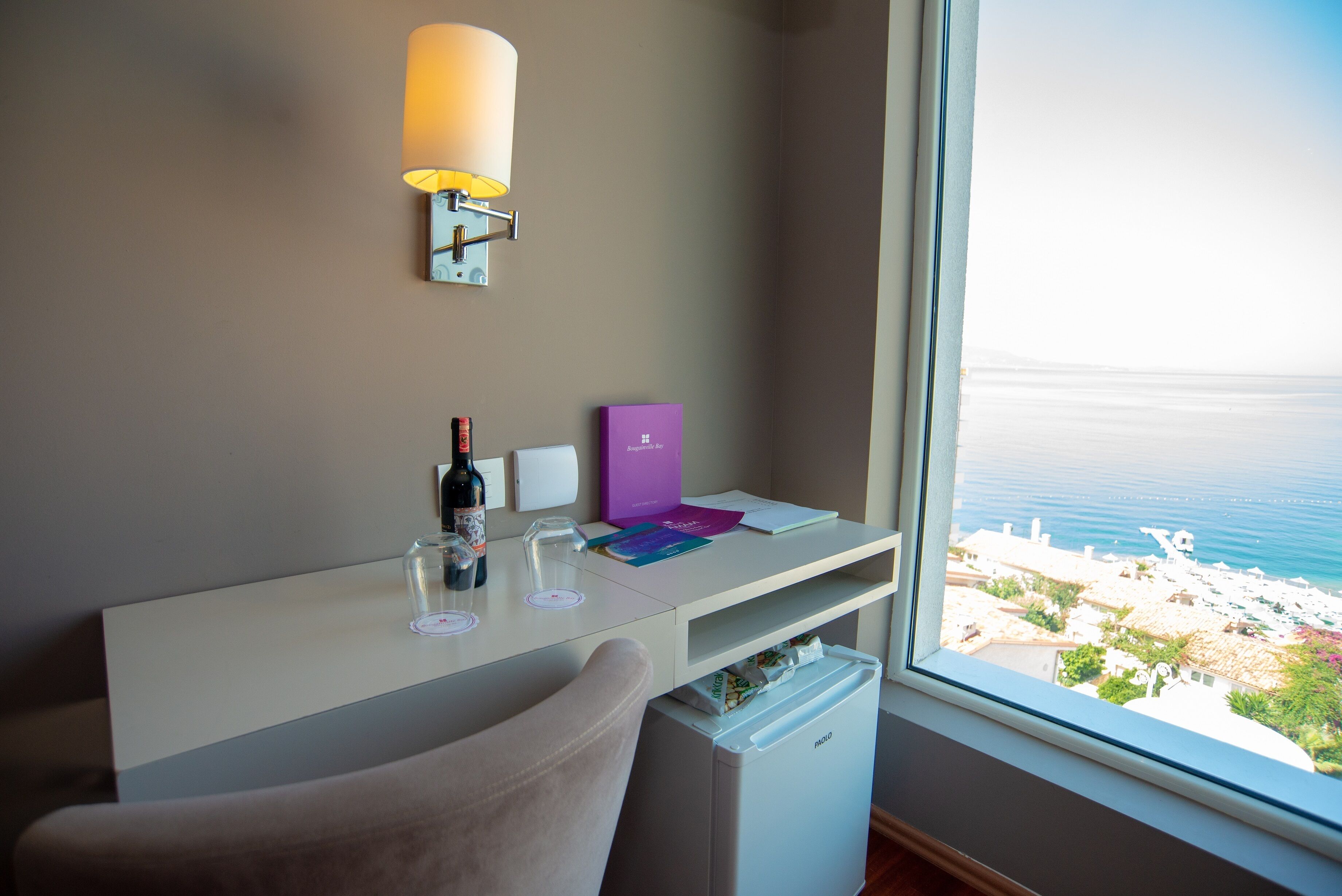 A hotel room with a desk, a chair, a lamp, a refrigerator, and a window with a view of the sea and buildings.