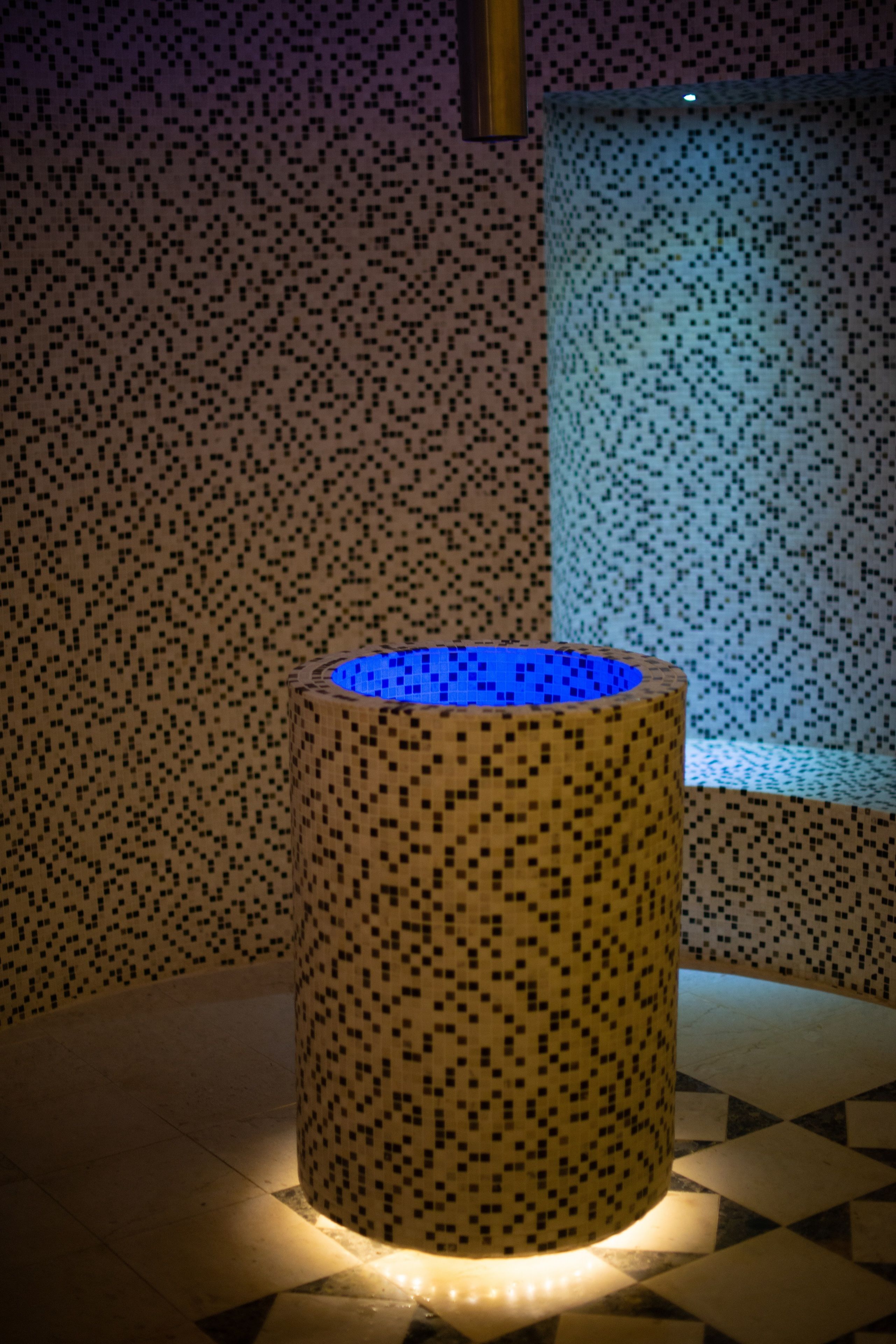 Un objet cylindrique orné d’un motif en mosaïque, éclairé par le bas, posé sur un sol carrelé à motifs géométriques.