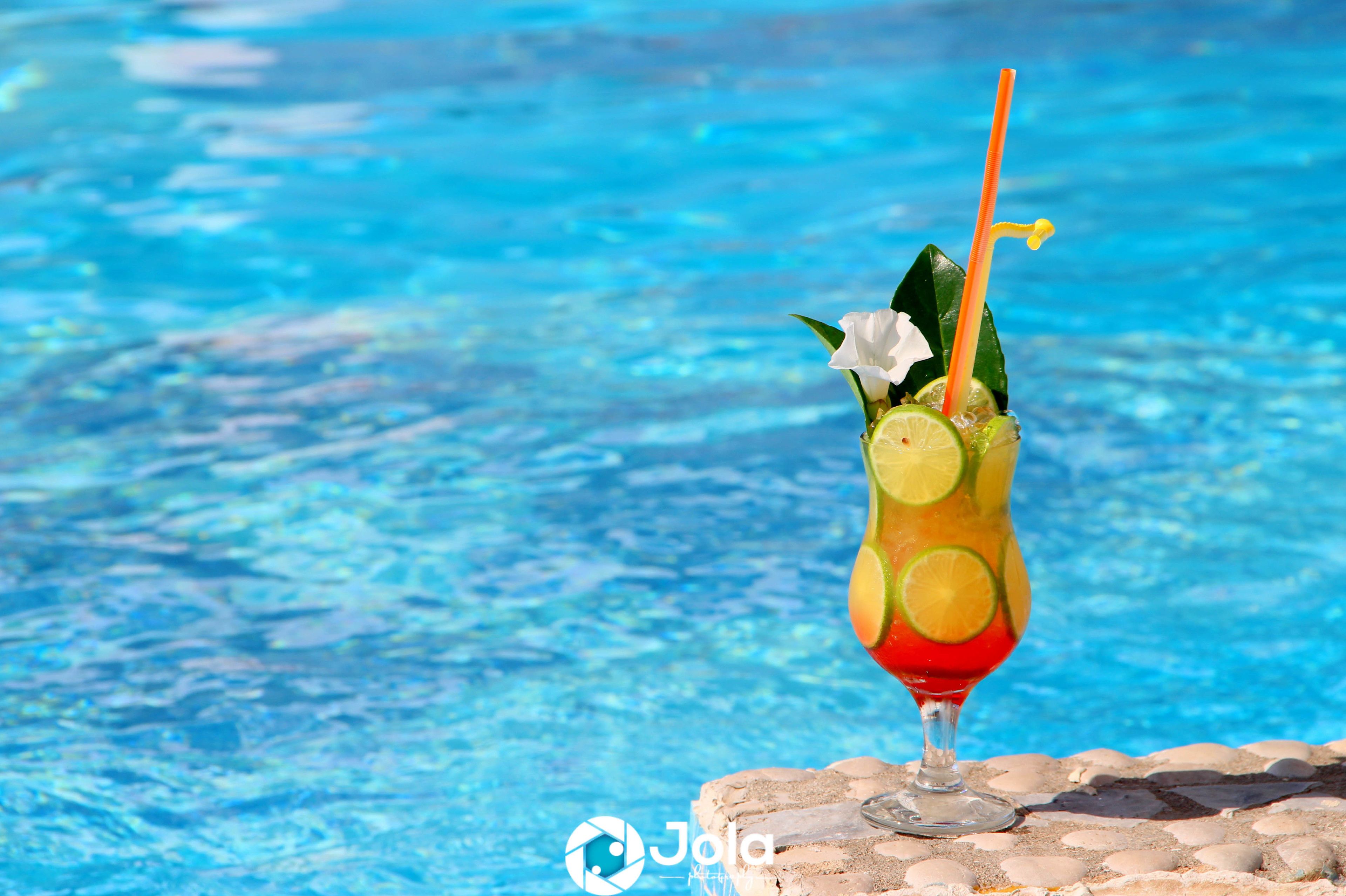 Ein Cocktail mit Limettenscheiben und einem Strohhalm in einem Glas, auf einer Steinfläche am Pool platziert.