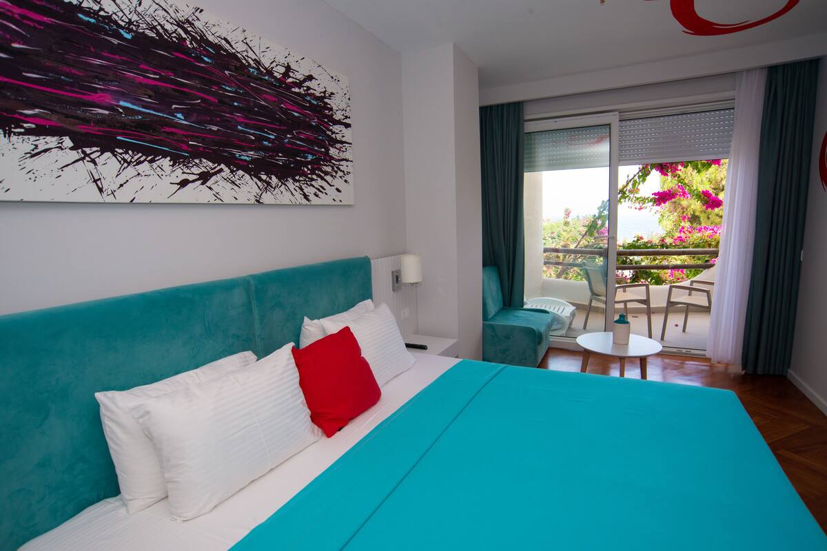 Een moderne hotelkamer met een groot bed, een blauwgroen hoofdeinde, witte beddengoed, een rood kussen, een kunstwerk aan de muur, een balkon met een tafel en stoelen, en uitzicht op groen en bloemen.