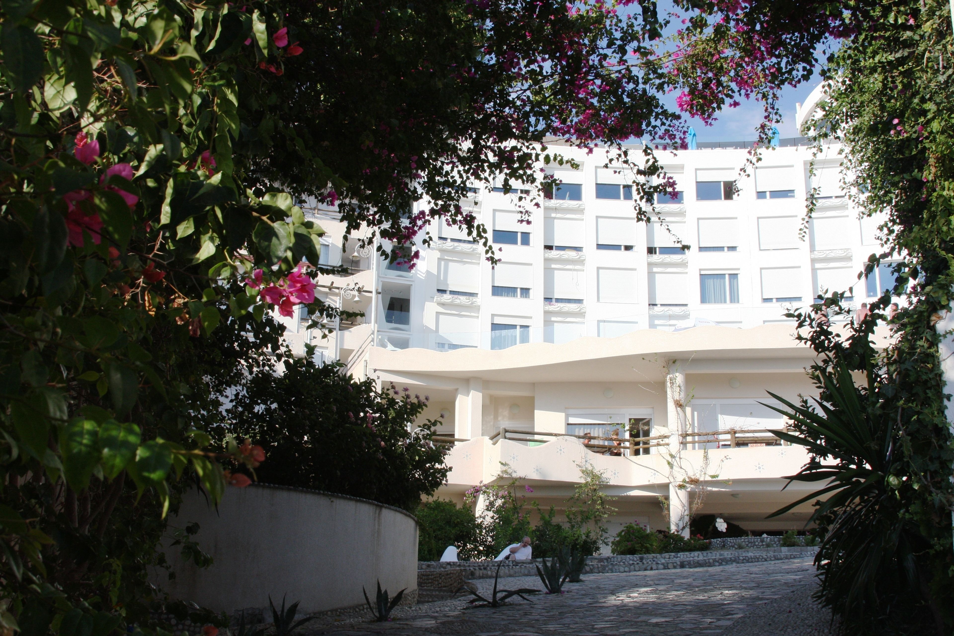 A modern building with a curved facade, surrounded by lush greenery and flowering plants.