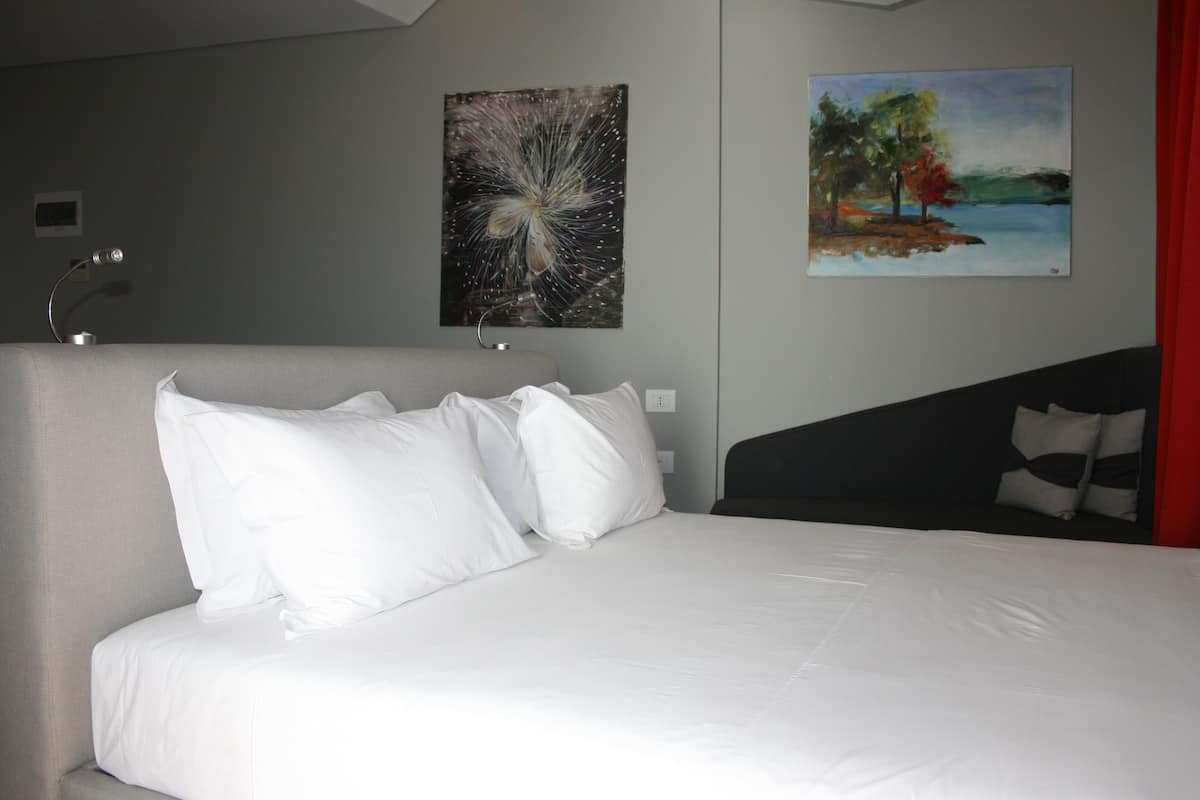 Een keurig opgemaakt bed met witte lakens, een moderne lamp en twee schilderijen aan de muur.