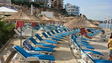 Ein Hotel am Strand mit Liegestühlen, Reetdachschirmen, einem Pool und einer felsigen Hügelkulisse im Hintergrund.
