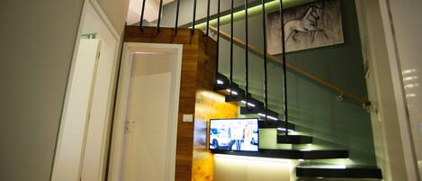 A staircase with wooden steps and a glass railing, leading to a landing with a flat-screen TV.