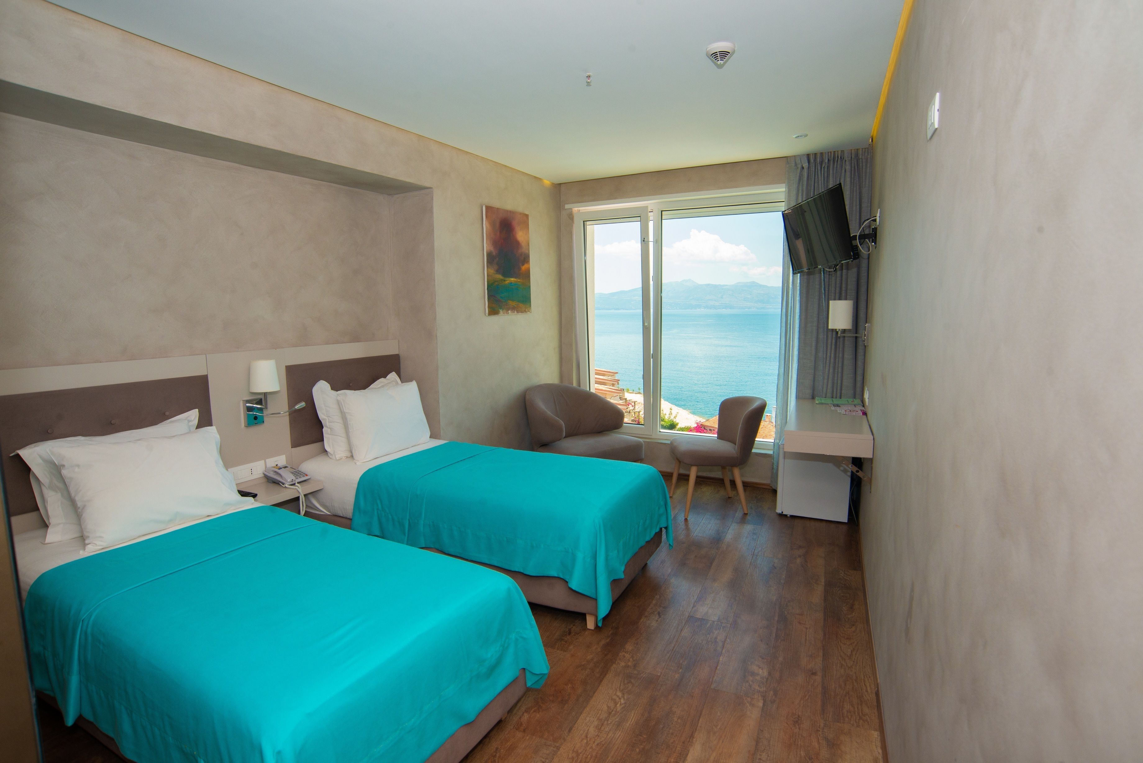 A hotel room with two beds, a desk, a chair, and a large window with a view of the sea.