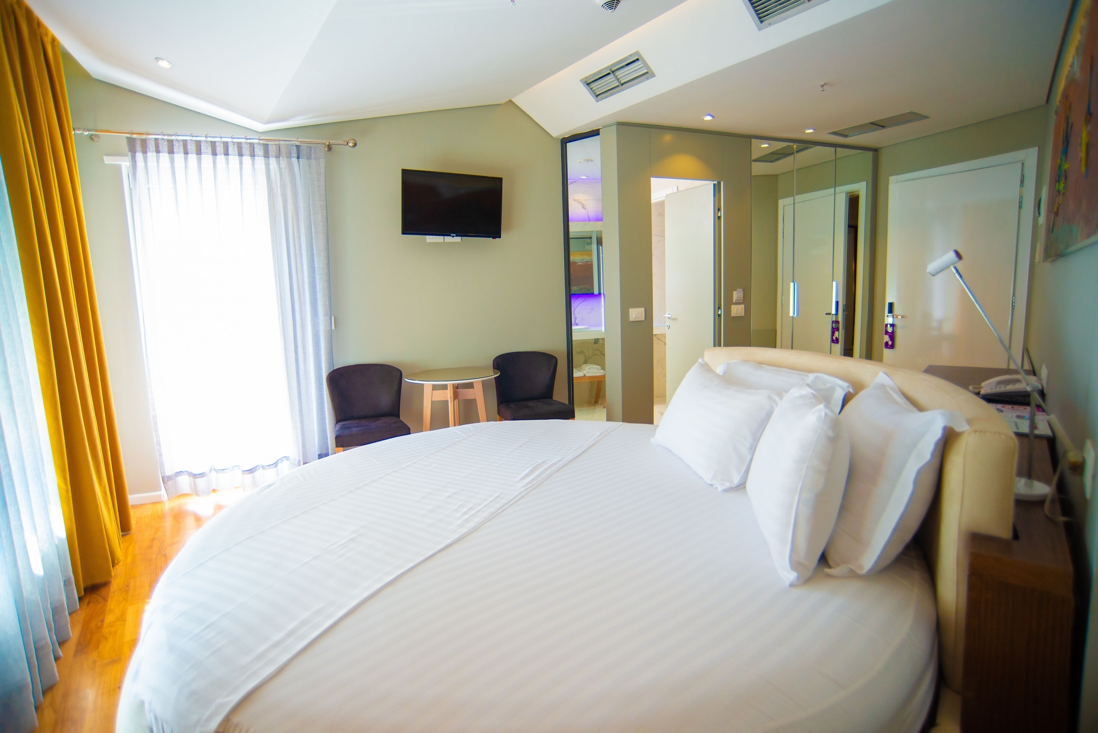 A large bed with white linens, a flat-screen TV mounted on the wall, a small round table, and a chair in a hotel room with a wooden floor and a large mirror.