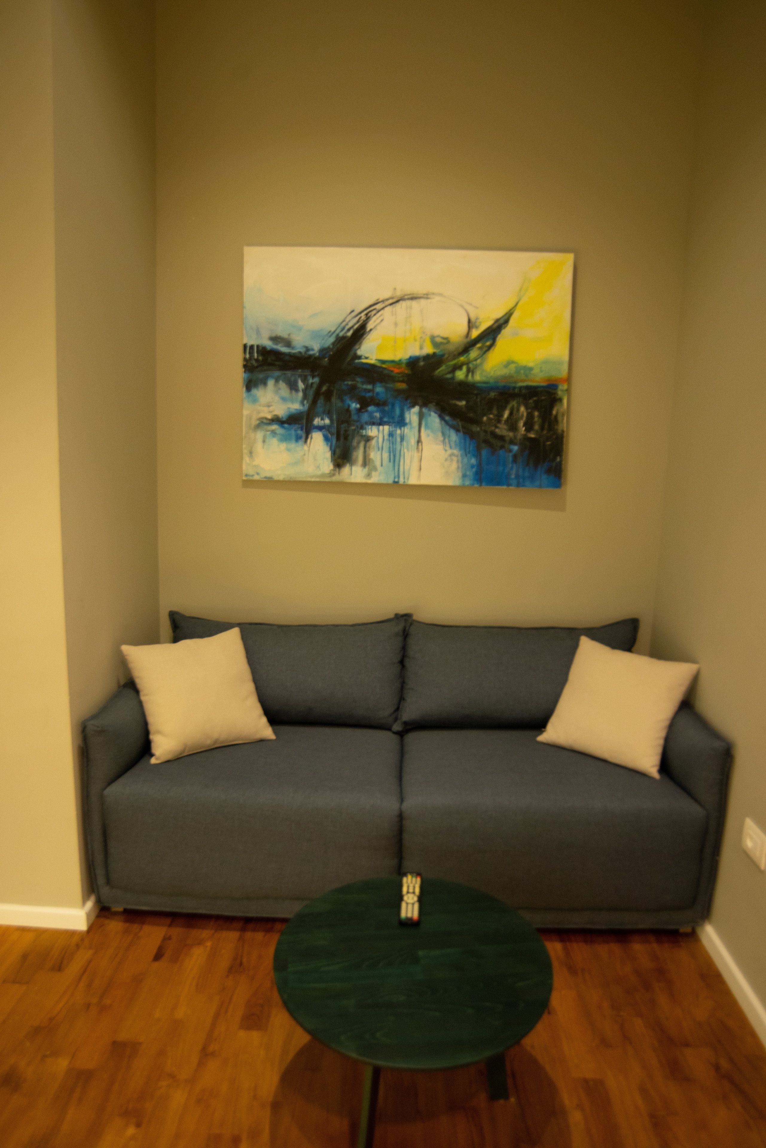 A gray sofa with two white cushions against a beige wall, a green round table with a remote control, and a painting above the sofa.