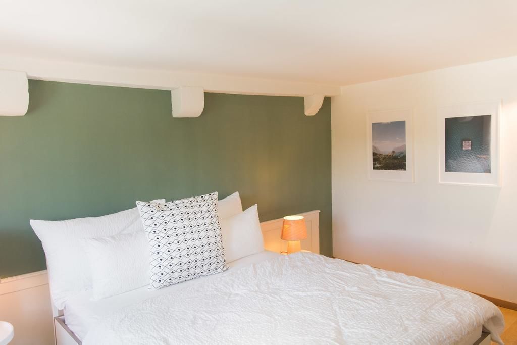 Una cama bien hecha con ropa blanca y almohadas estampadas, una lámpara de noche y dos cuadros en la pared.