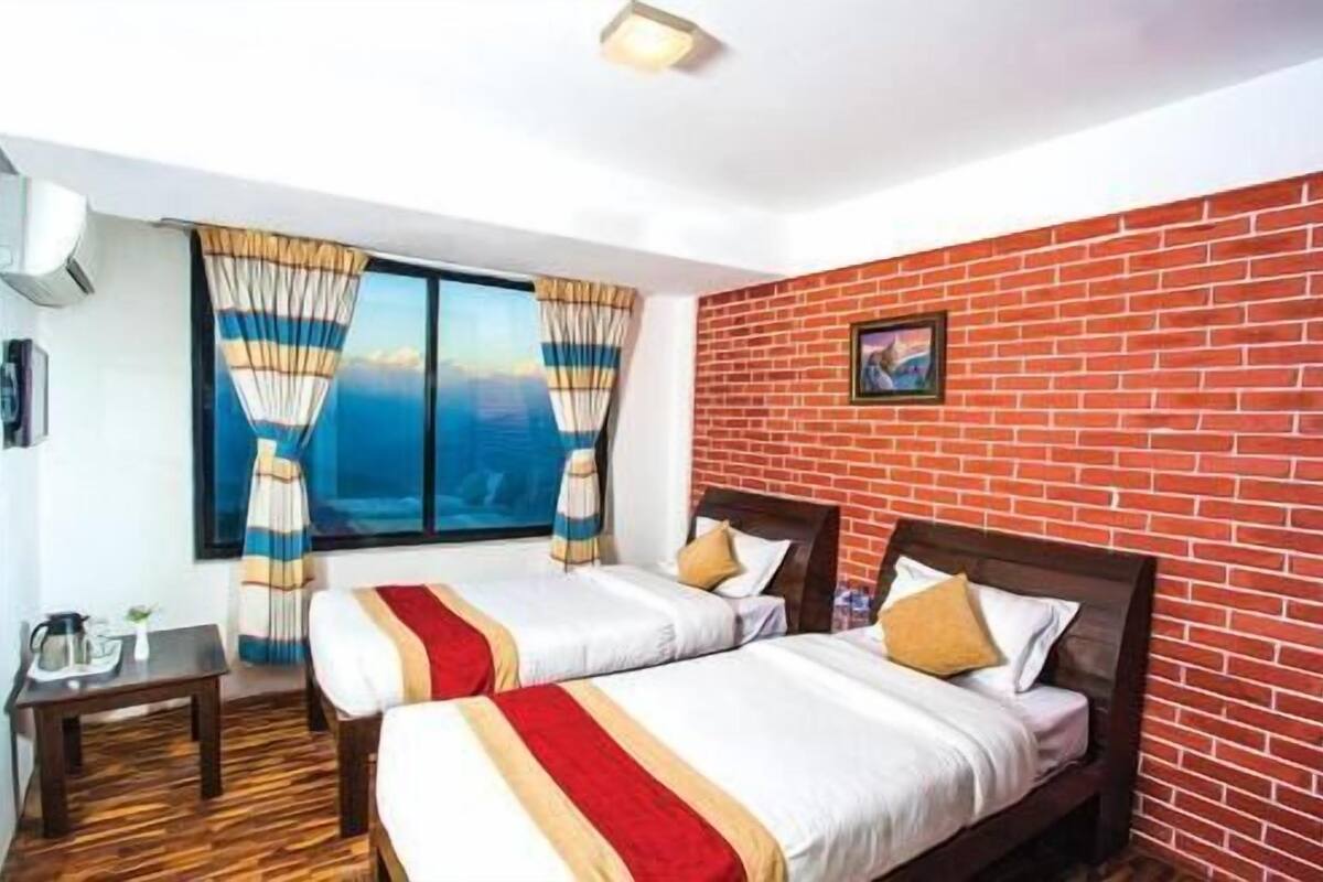 A hotel room with two beds, a brick wall, a window with curtains, a small table, and a picture on the wall.
