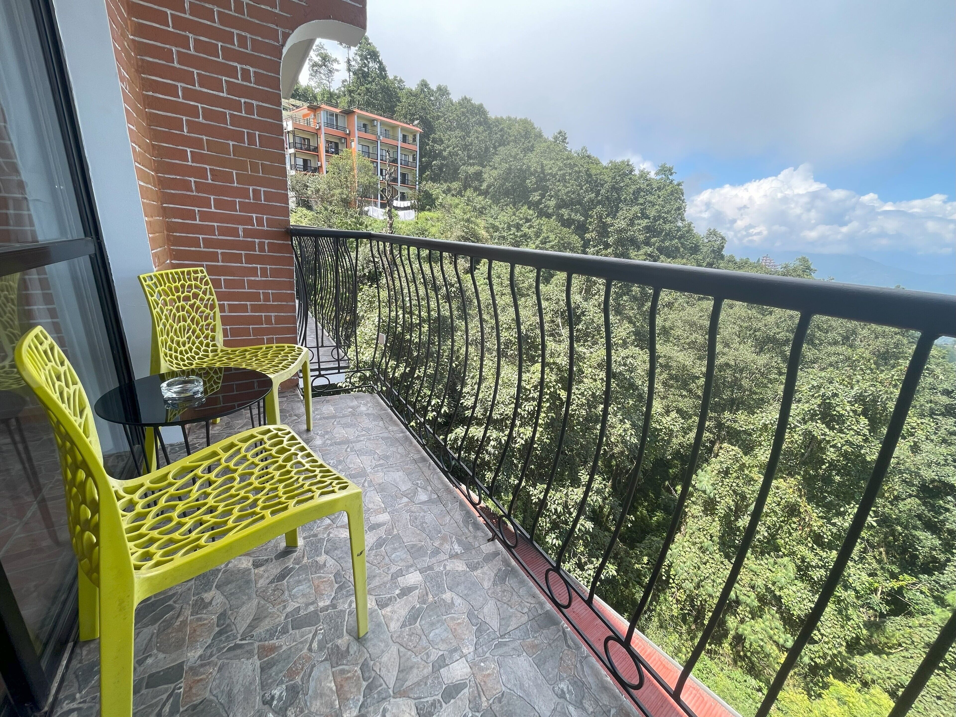 A balcony with yellow chairs and a table, overlooking a forested area and a building in the background.