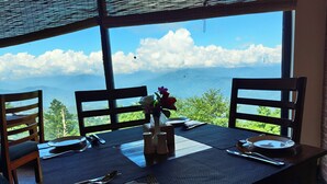 A dining table with chairs, a vase with flowers, and a view of mountains and clouds through large windows. - Hotel Himalayan Villa - Nagarkot (Shankharapur)