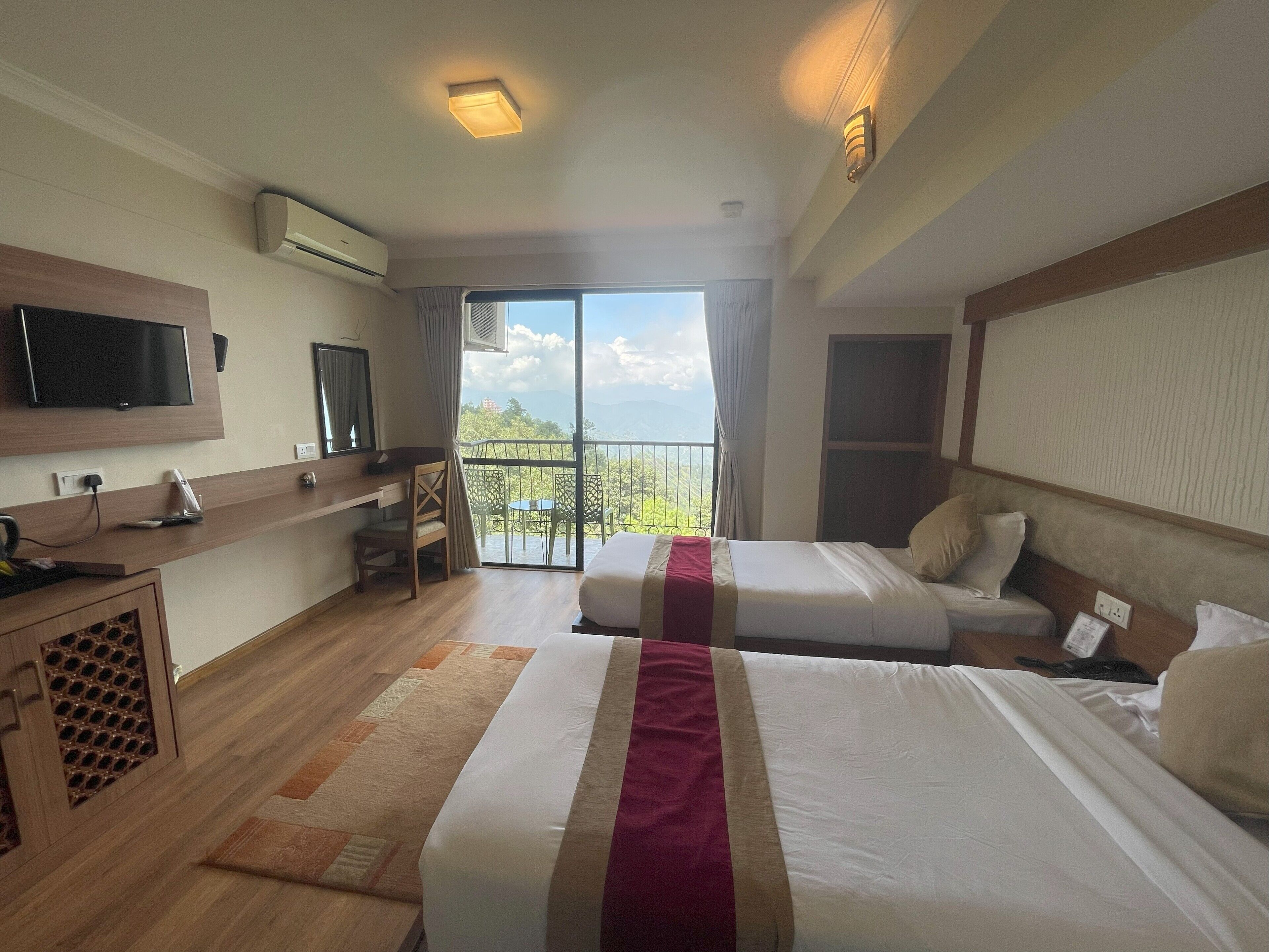 A hotel room with two beds, a desk, a TV, and a balcony with a view.