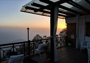 A wooden deck with outdoor seating, a pergola, and a view of the sunset over a mountainous landscape. - Hotel Himalayan Villa - Nagarkot (Shankharapur)