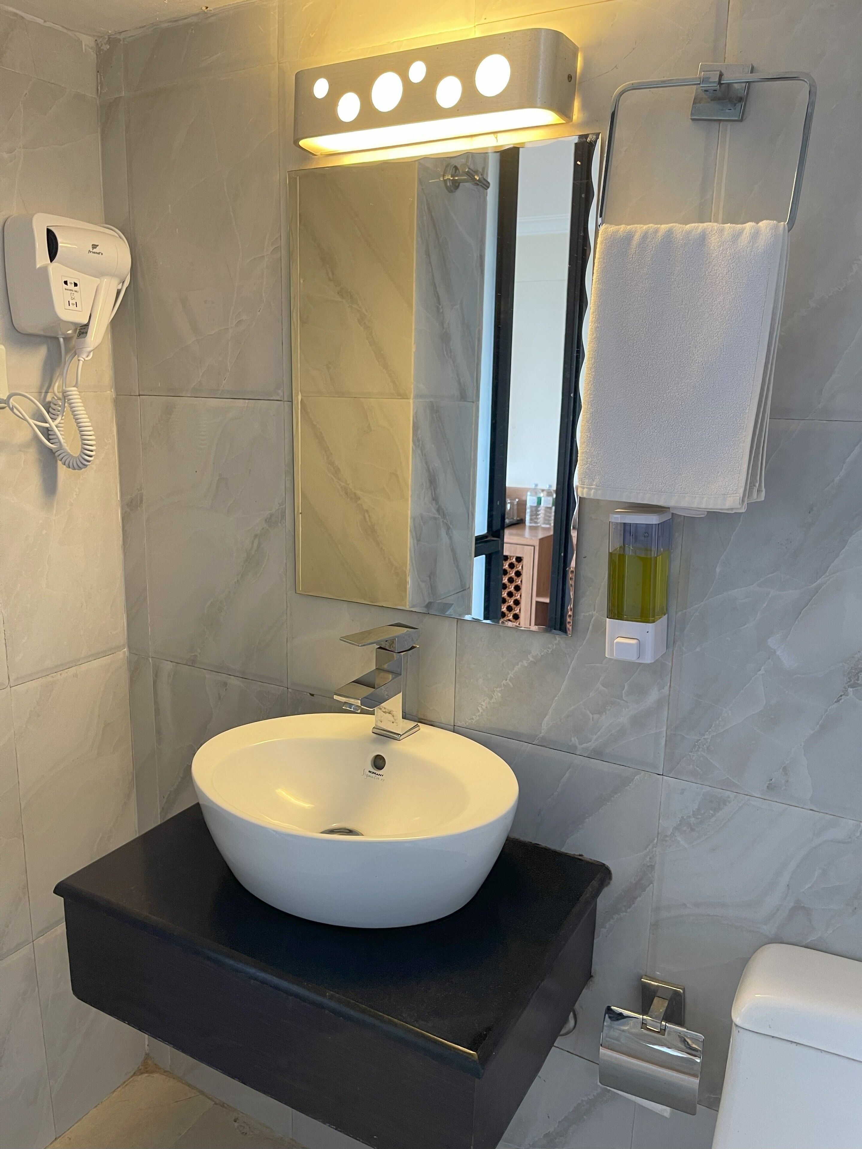 A bathroom with a white sink, a mirror with lights, a towel rack, and a hand dryer.