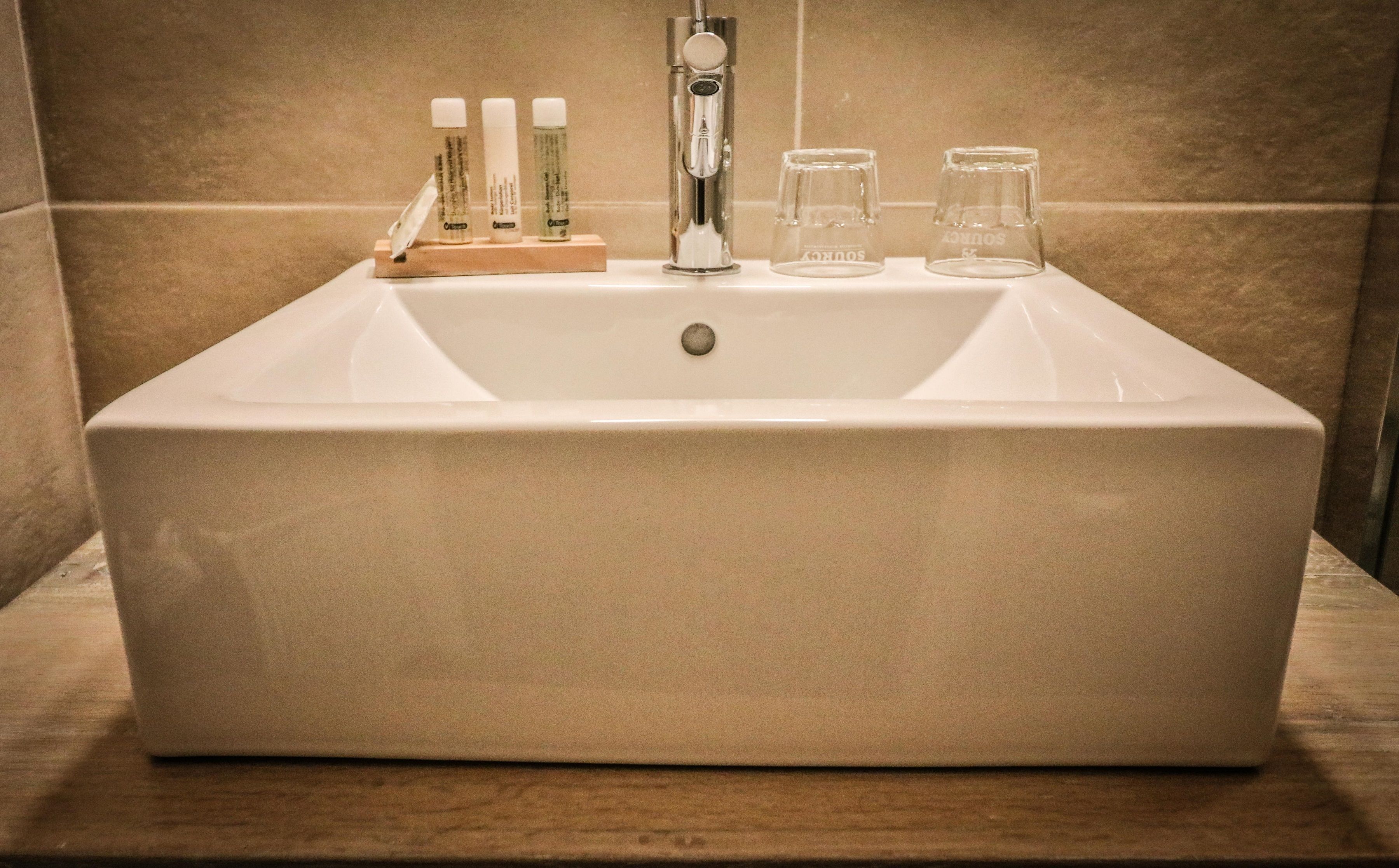 Un lavabo de baño rectangular con un grifo moderno, tres botellas de jabón líquido y dos vasos de cristal.