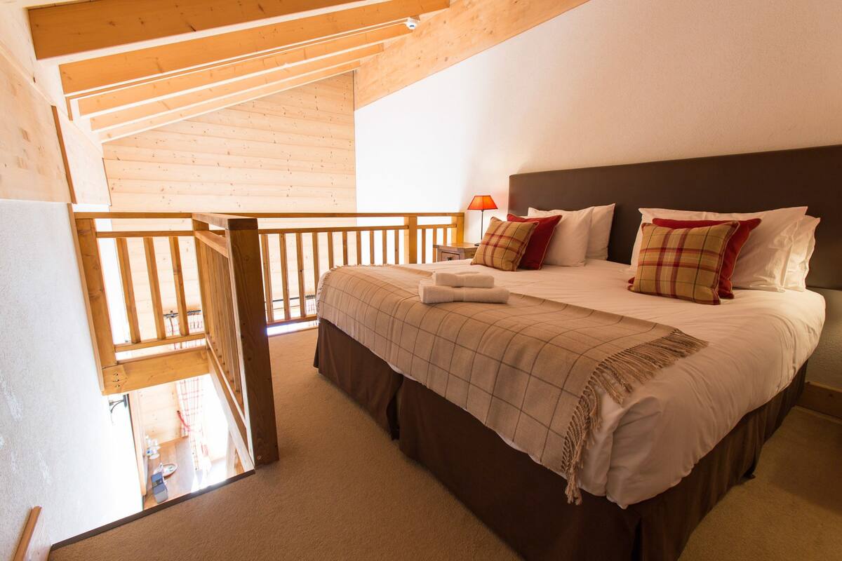 A bedroom with a wooden ceiling, a bed with a plaid bedspread, and a nightstand with a lamp.
