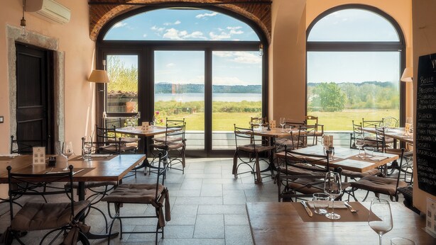 Un restaurante con mesas y sillas, amplios ventanales arqueados y vistas a un lago y follaje.