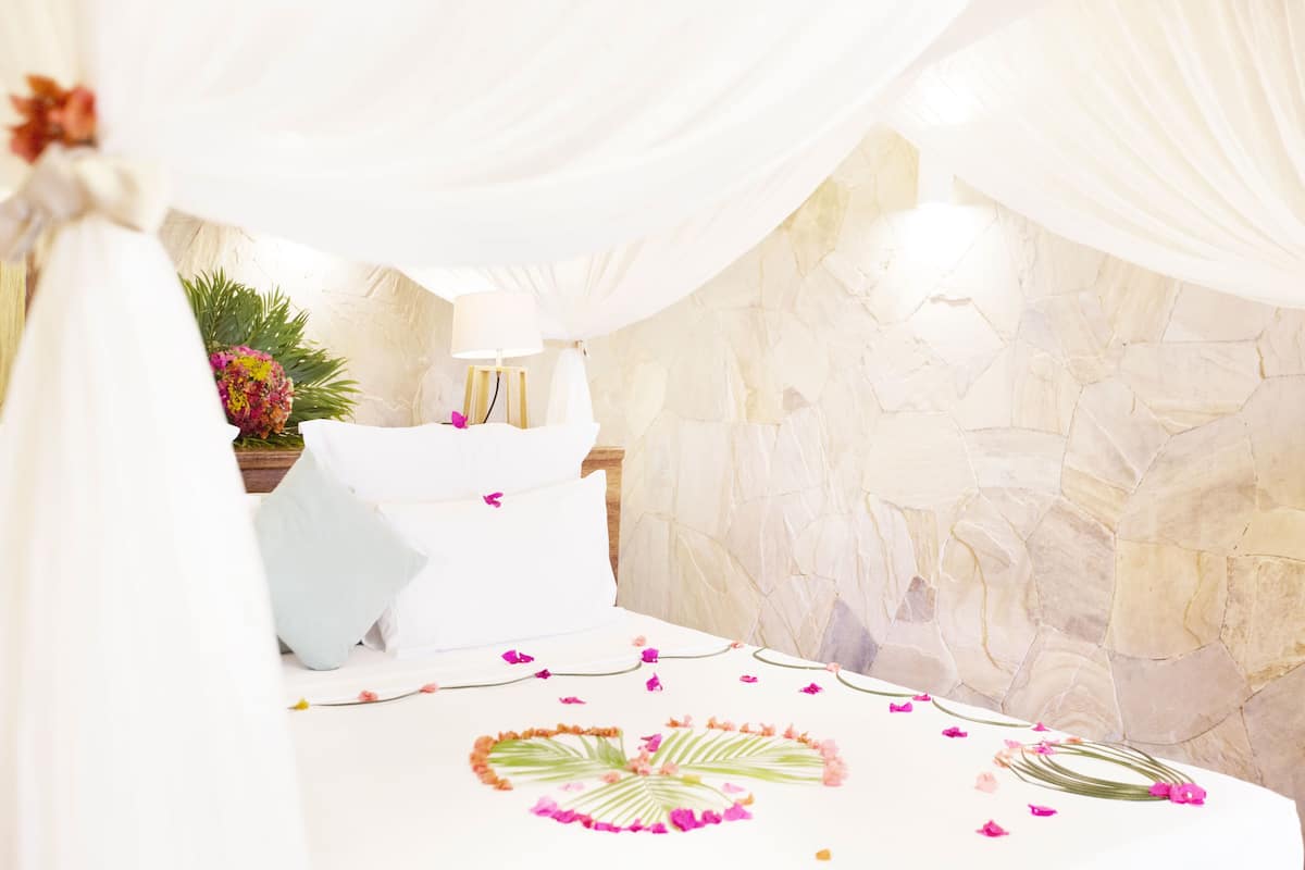 A bed with white linens, a stone wall, and a lamp.