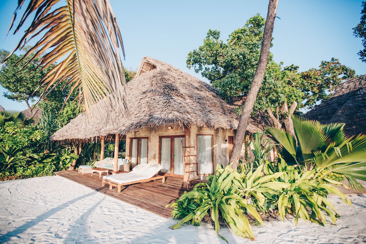 A beachside hut with a thatched roof, wooden deck, and two lounge chairs.