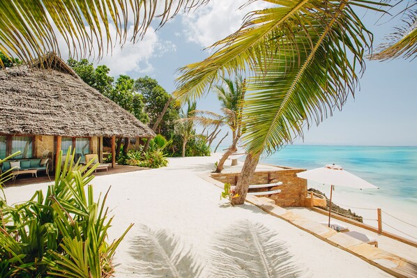 A beach with a thatched-roof hut, lounge chairs, and a palm tree. - Tulia Zanzibar Unique Beach Resort (Pongwe)