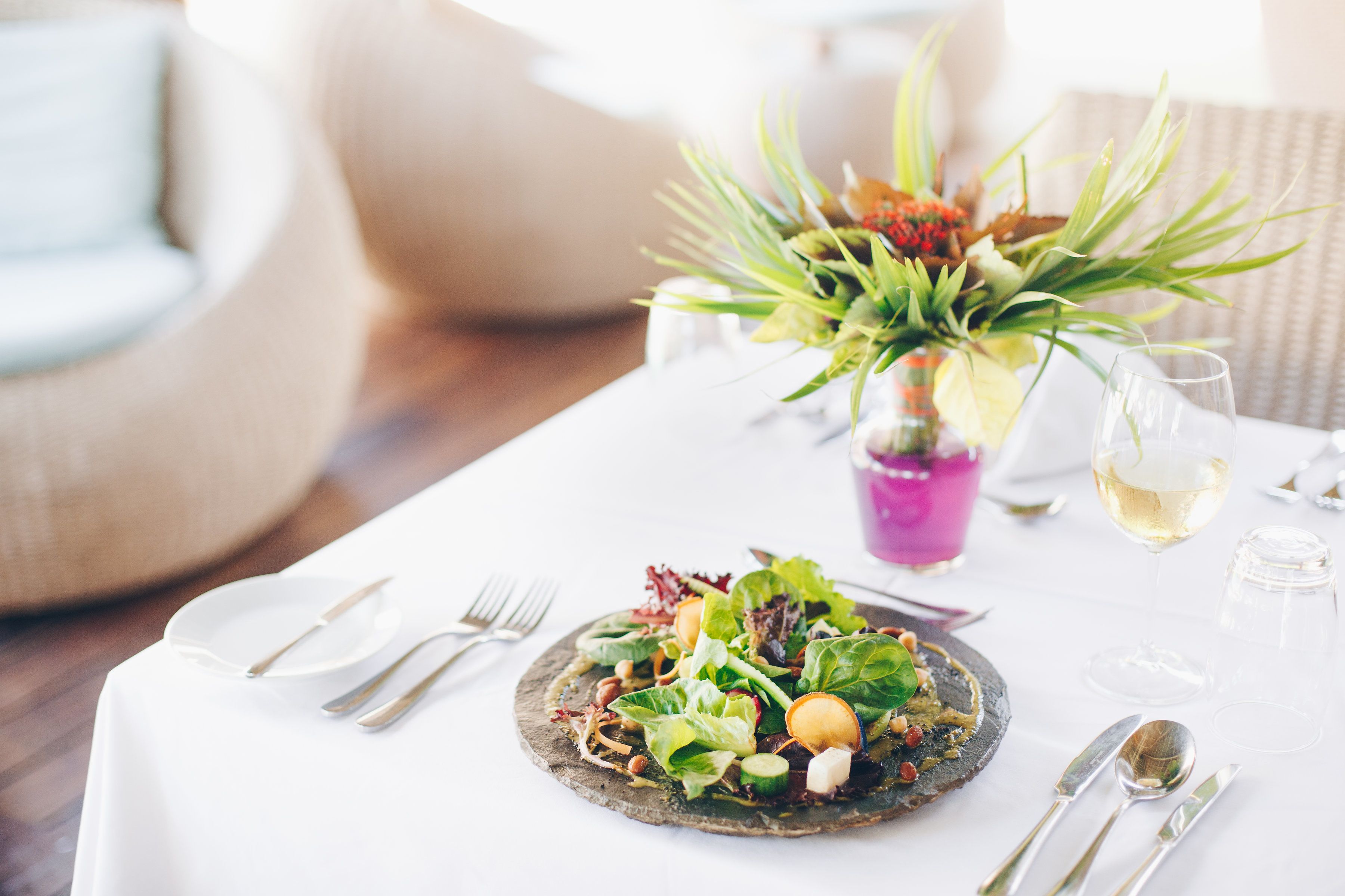 Uma mesa de jantar com salada, talheres e um copo de vinho branco.