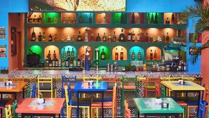 A colorful bar with a variety of bottles and a bartender. There are multiple tables and chairs with colorful upholstery. - Scarlet Lodge (Lagos)