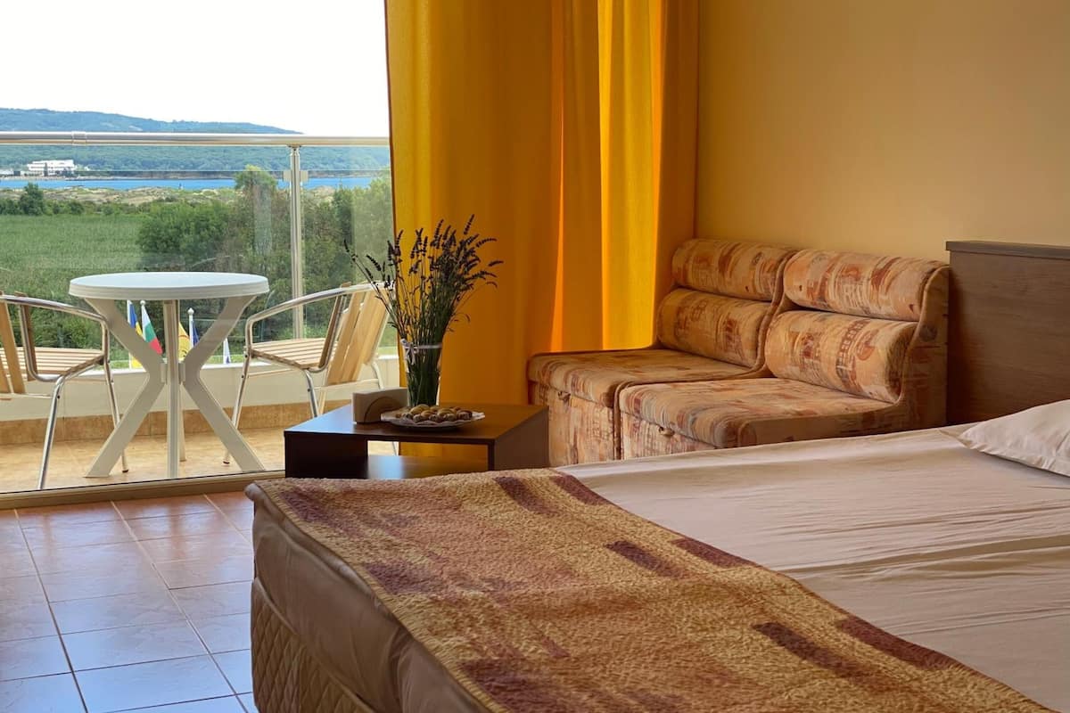 Ein Hotelzimmer mit Bett, Sofa, einem kleinen Tisch mit Blumenvase und einem Balkon mit Tisch und Stühlen.